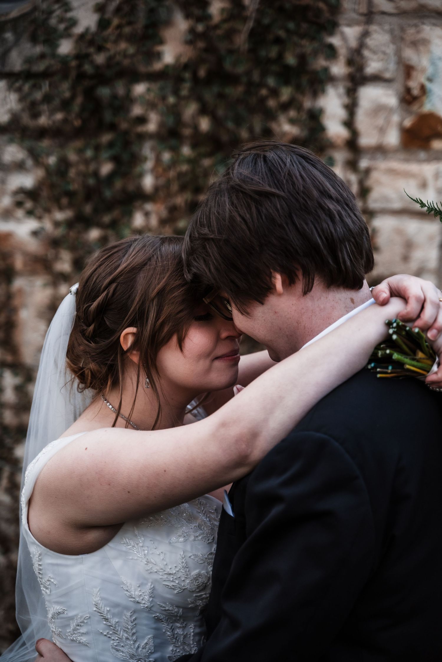 An intimate moment between newlyweds as they embrace against a vine-covered stone wall backdrop.