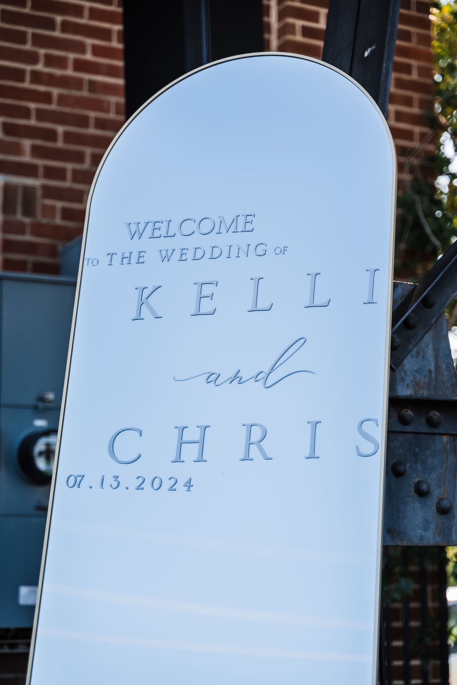 Modern white welcome sign for Kelli and Chris's wedding displayed against a brick building.