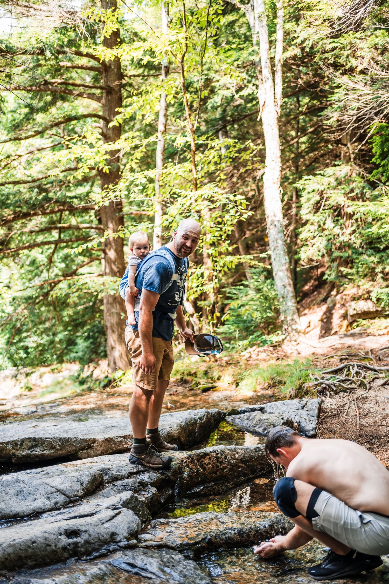 Two hikers explore a rocky stream area surrounded by lush forest on a sunny day.