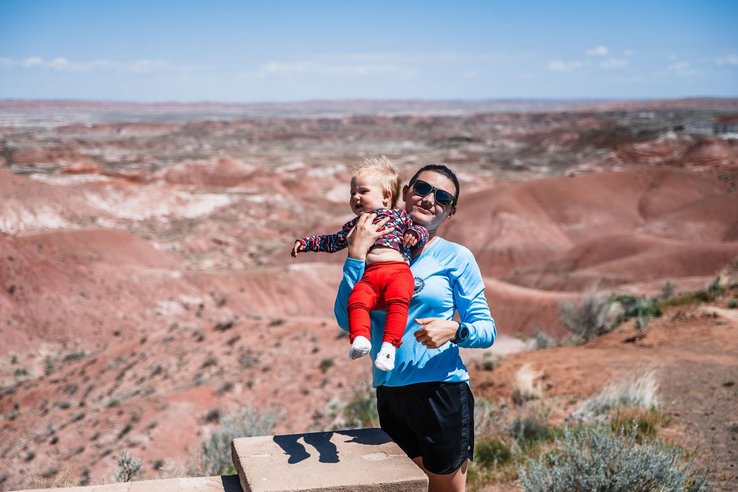 Hiking along desert trail while carrying small child with red rock formations in background.
