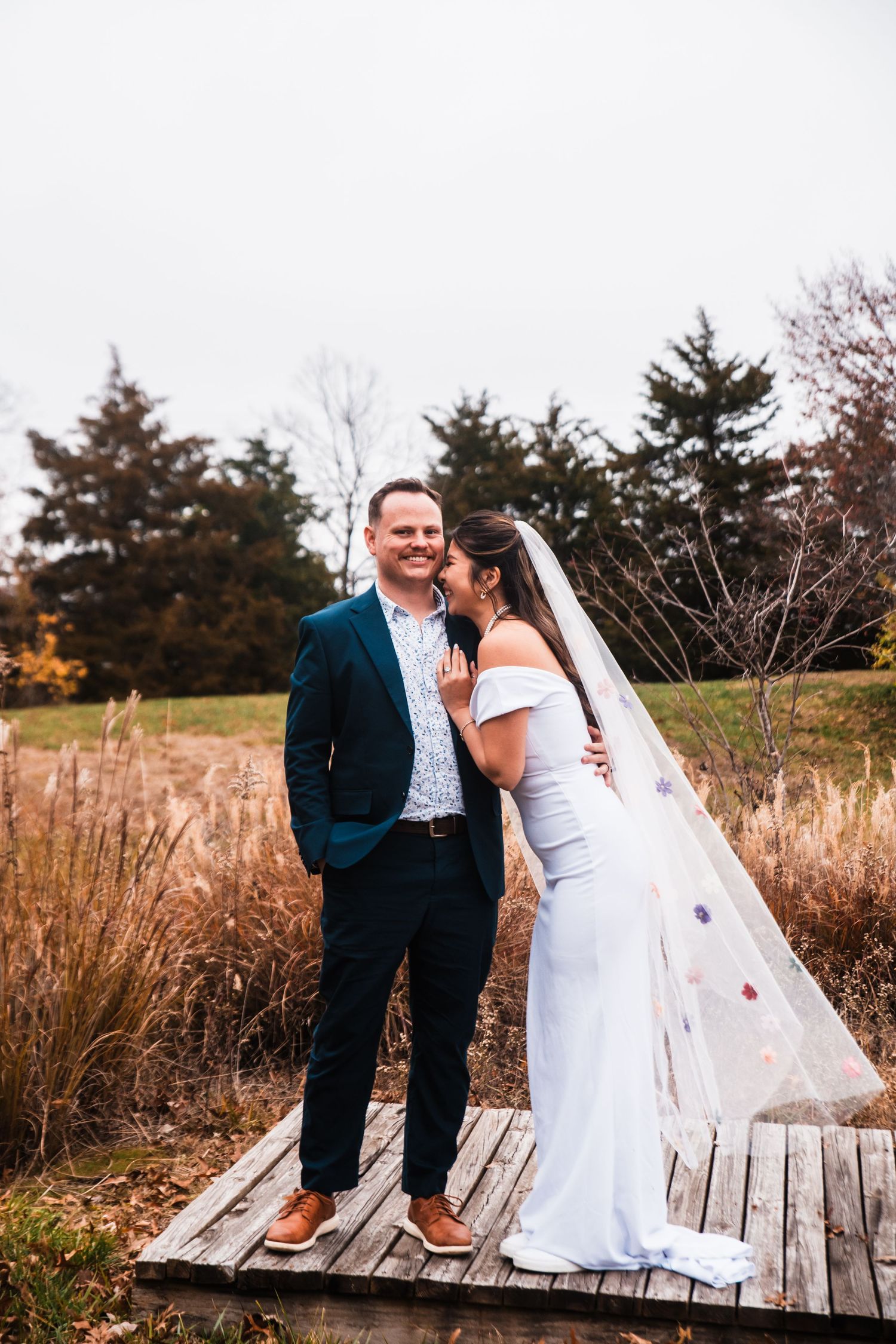A couple stands together on a wooden deck surrounded by autumn foliage.
