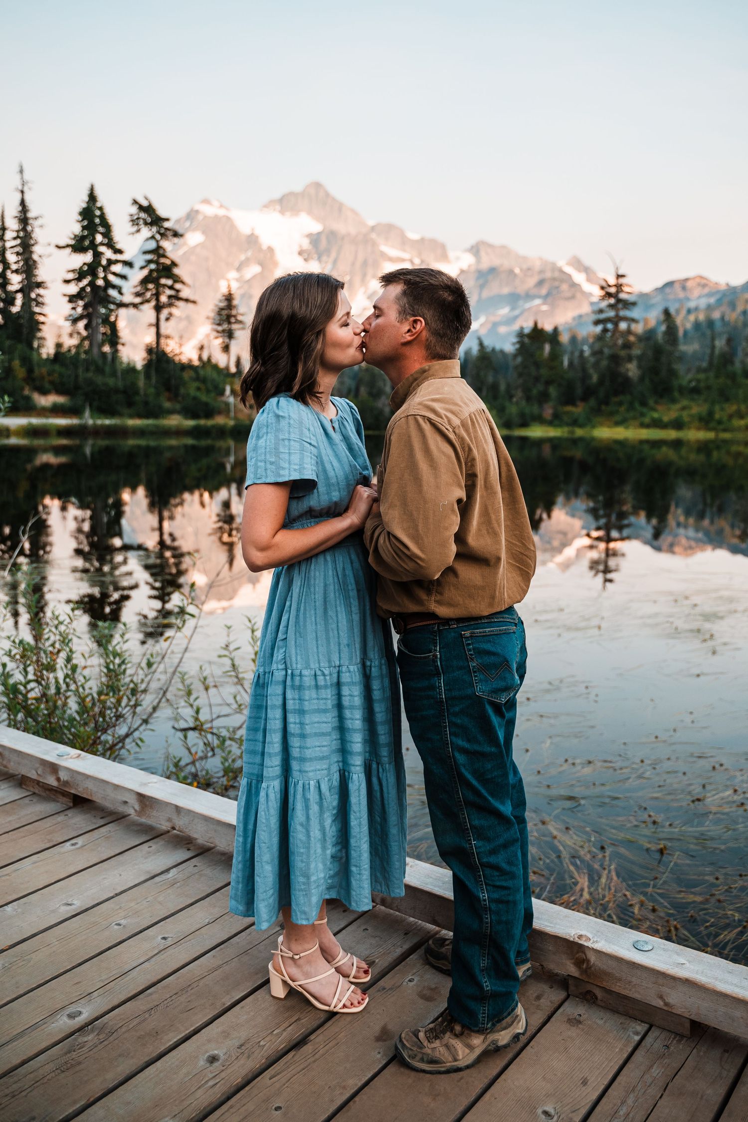 A couple shares a romantic moment on a wooden dock overlooking a mountain lake at sunset.