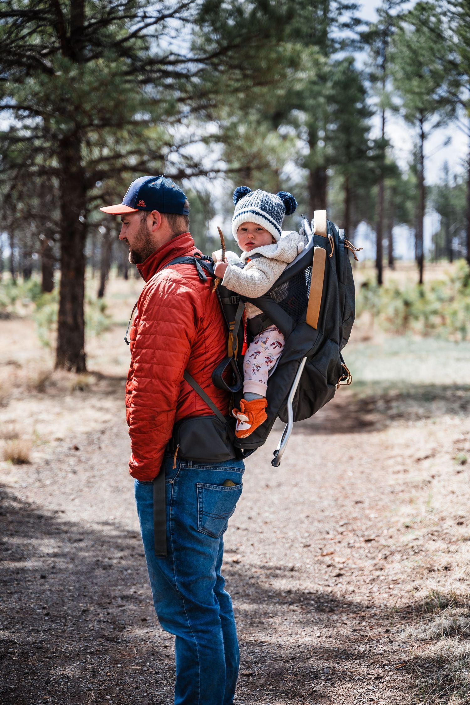 A hiker in a red jacket carries a small passenger in a backpack carrier through a pine forest.