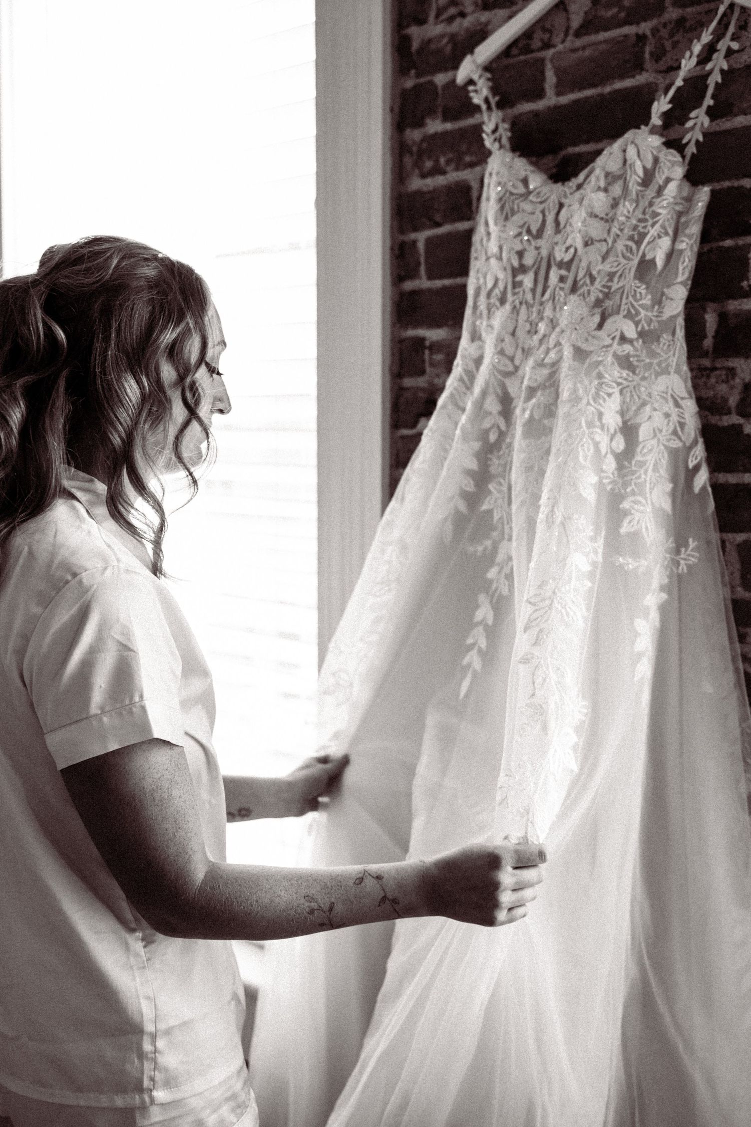 A wedding dress hangs in front of a window while someone in white examines the delicate details.