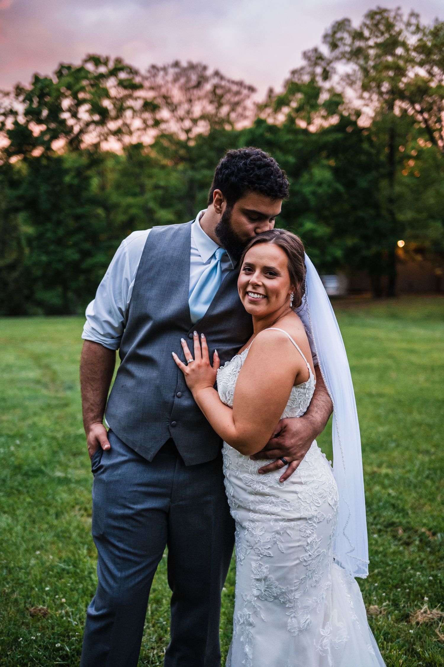 Newlywed couple embracing outdoors at sunset with bride in lace dress and groom in grey vest.