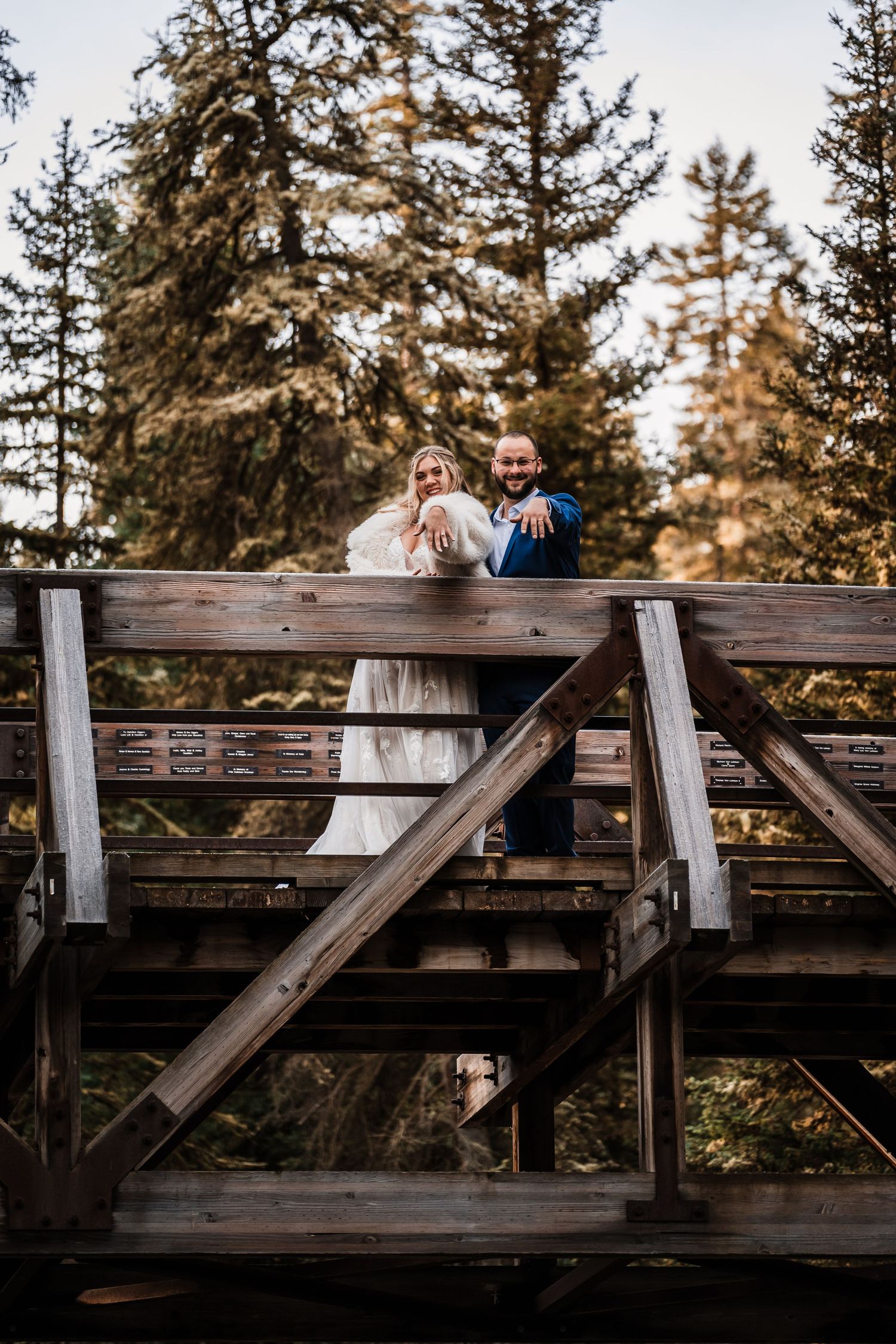 Couple in wedding attire on rustic wooden bridge surrounded by evergreen trees.