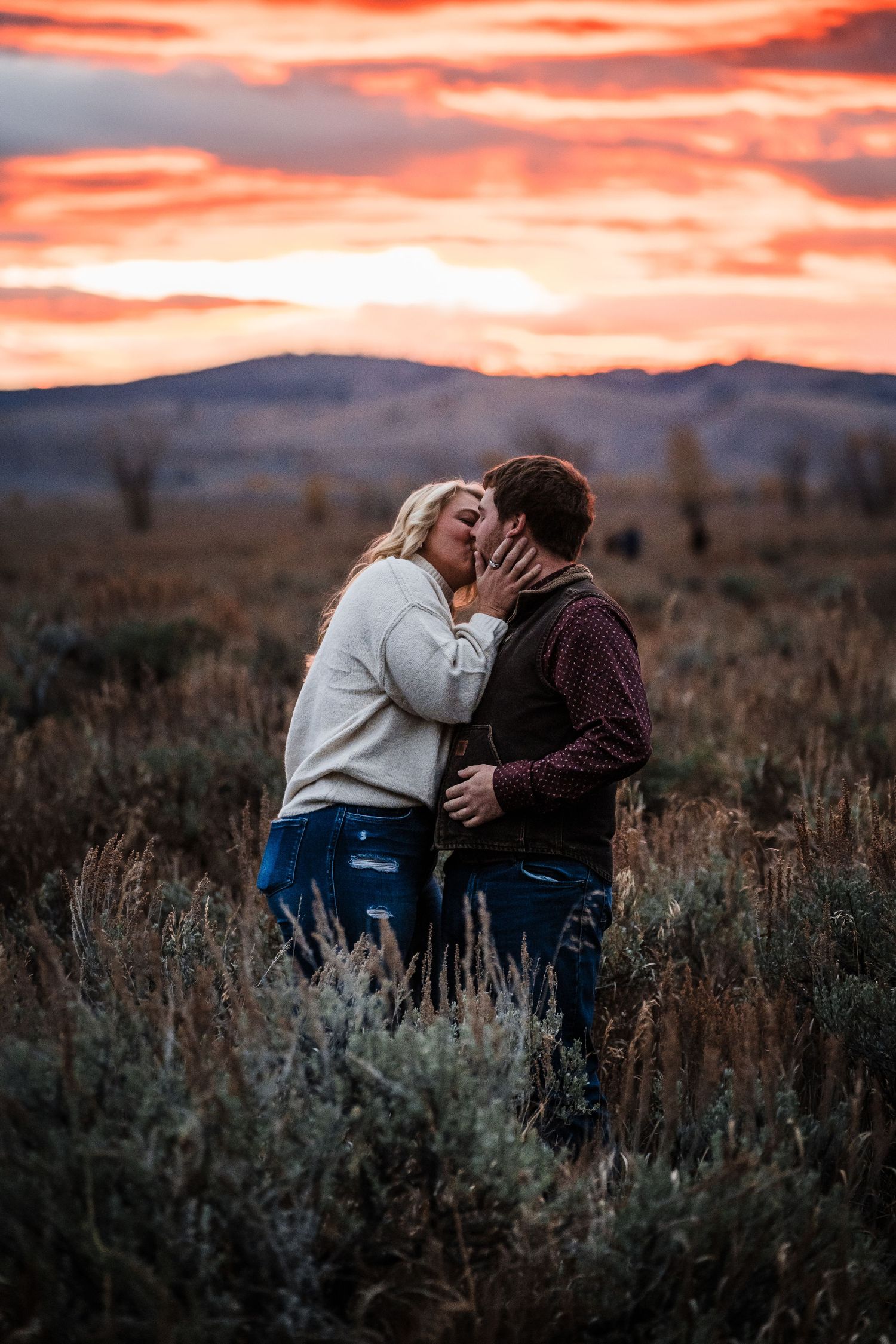 A couple shares a romantic moment in a field of sagebrush during a vibrant sunset in the mountains.