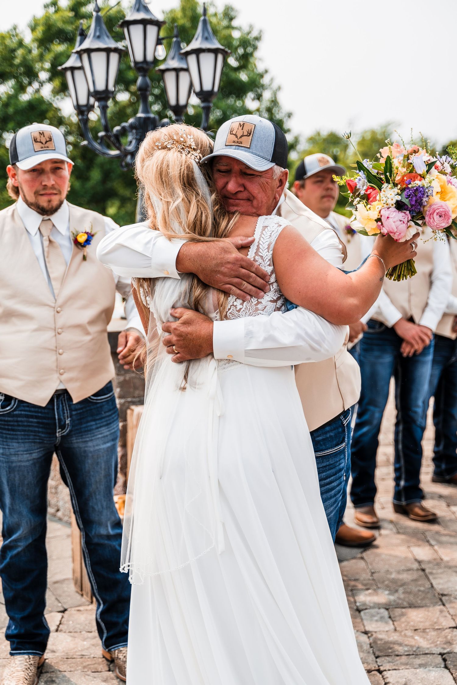 Wedding party members embrace in a line on a stone patio with decorative lanterns visible.