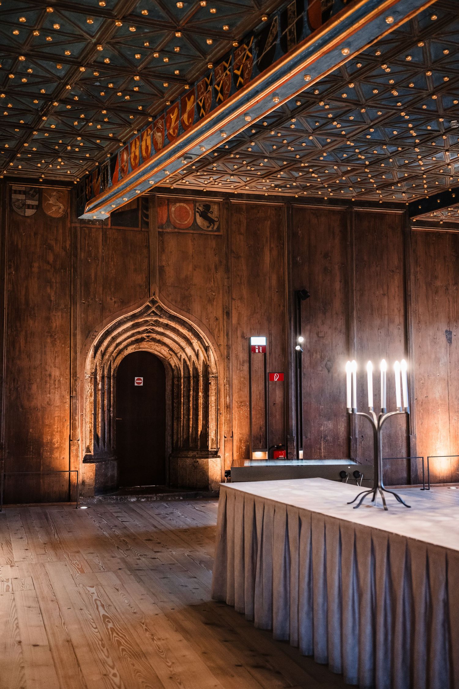 Medieval-style hall with wooden panels and ornate ceiling featuring candelabras and gothic arches in warm lighting.