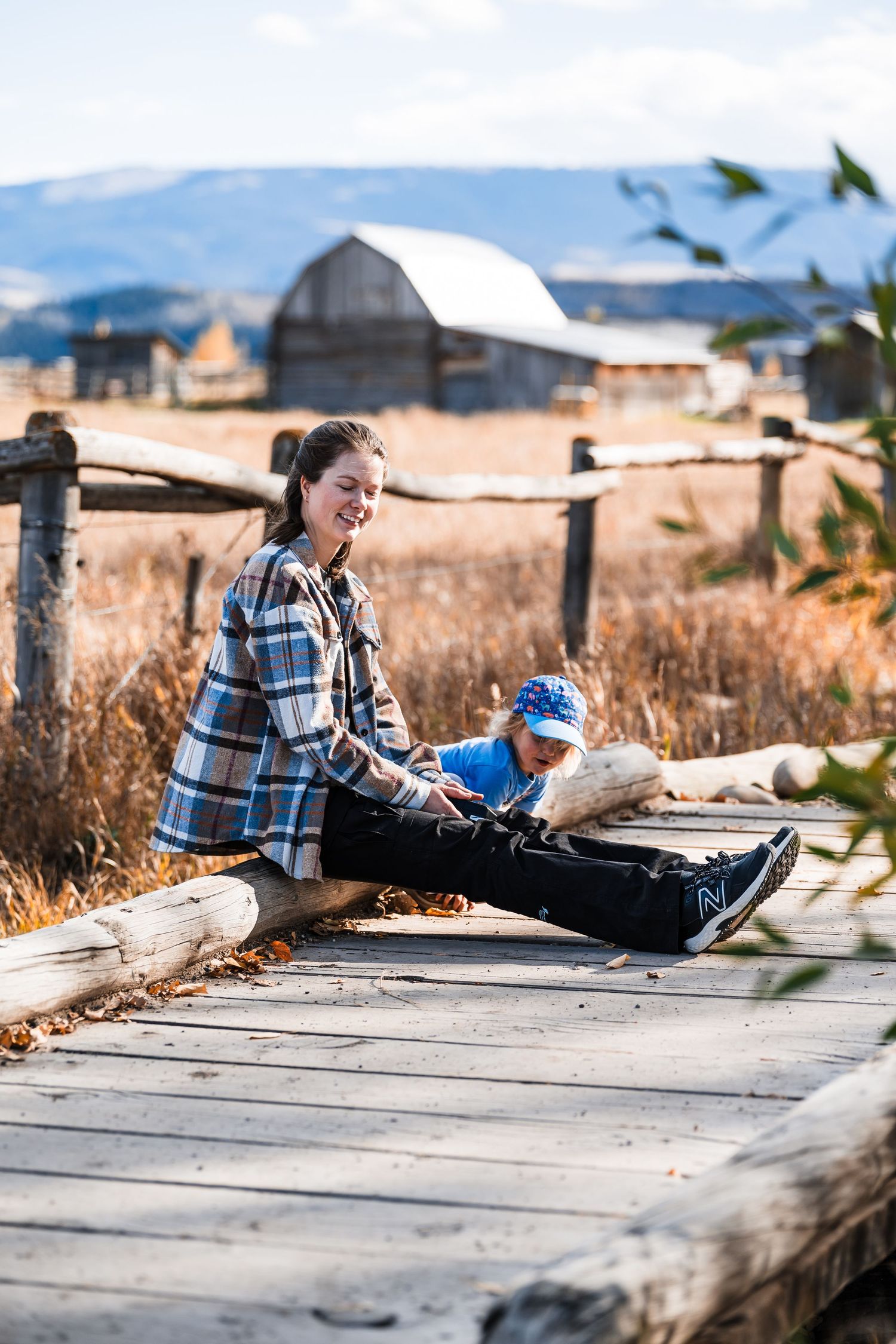 Someone in a plaid flannel shirt sits on a wooden boardwalk with mountains and a rustic barn in the background.
