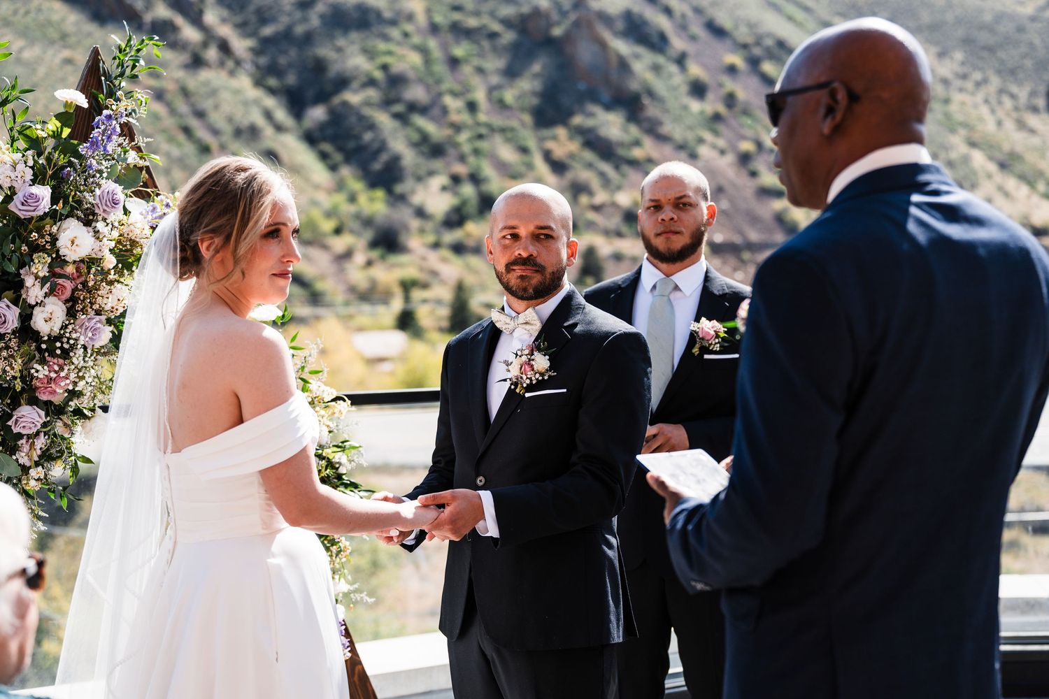 Outdoor wedding ceremony with bride and groom exchanging vows before an officiant, floral arch backdrop, mountain scenery.