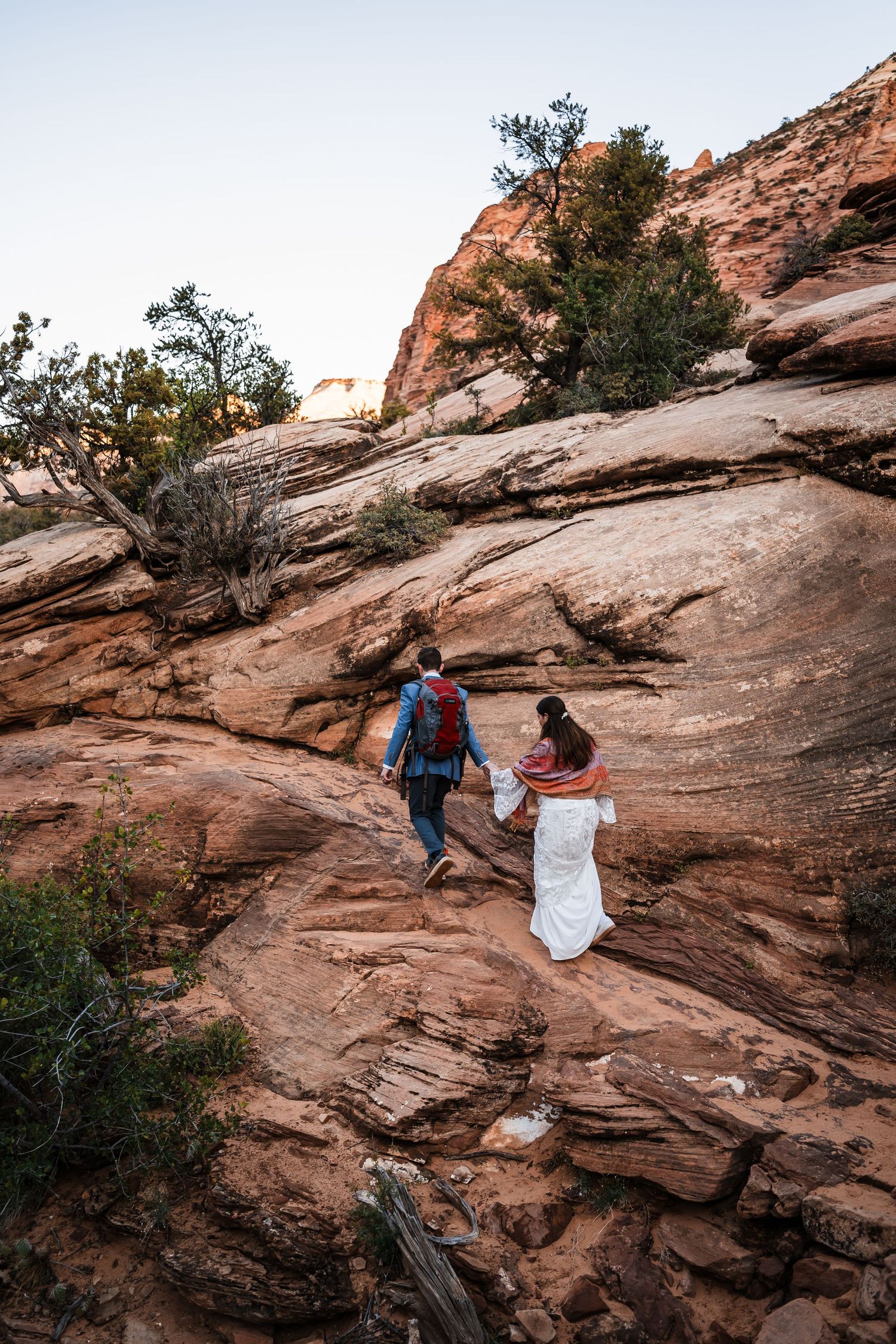 Bride and groom in wedding attire hiking up red rock formations at Zion National Park at sunset.