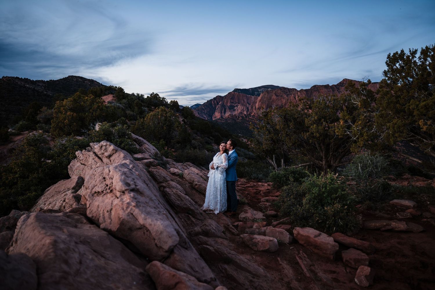 Bride and groom embracing among red rocks at twilight with dramatic Sedona mountain backdrop.