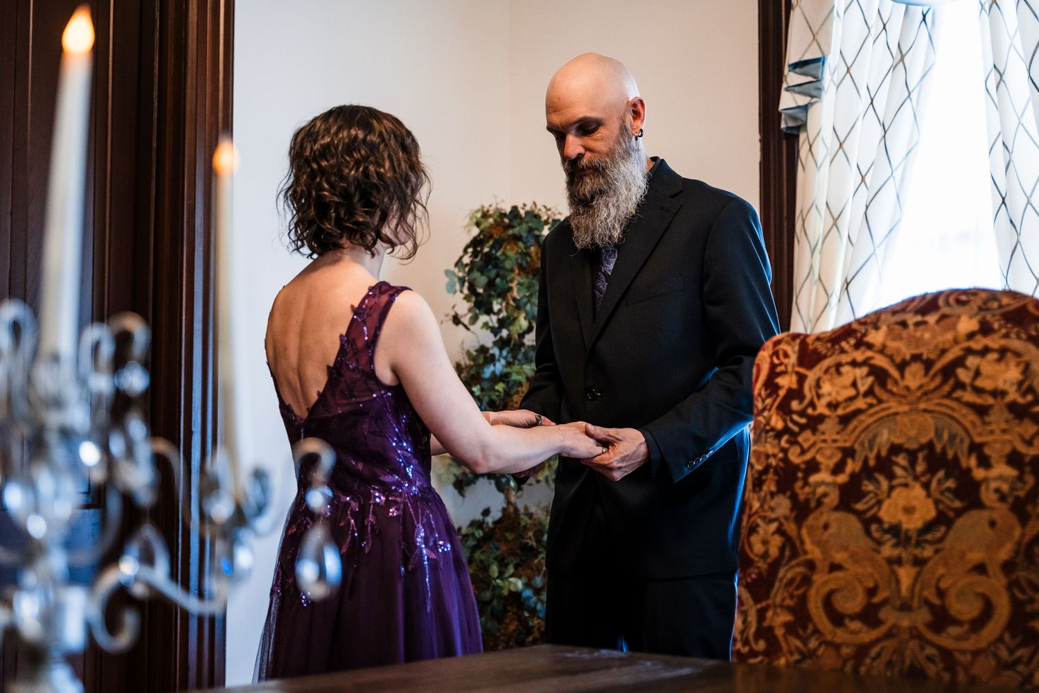 Bald groom with gray beard exchanges rings with bride in purple dress during intimate wedding ceremony indoors.