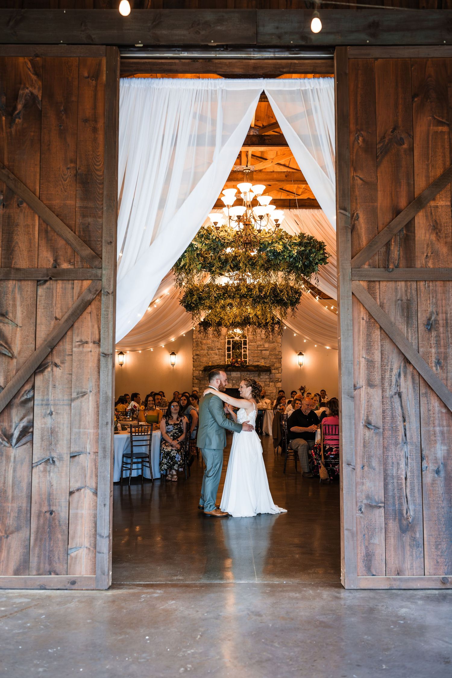 Bride and groom share first dance framed by rustic barn doors with glowing chandelier and string lights.