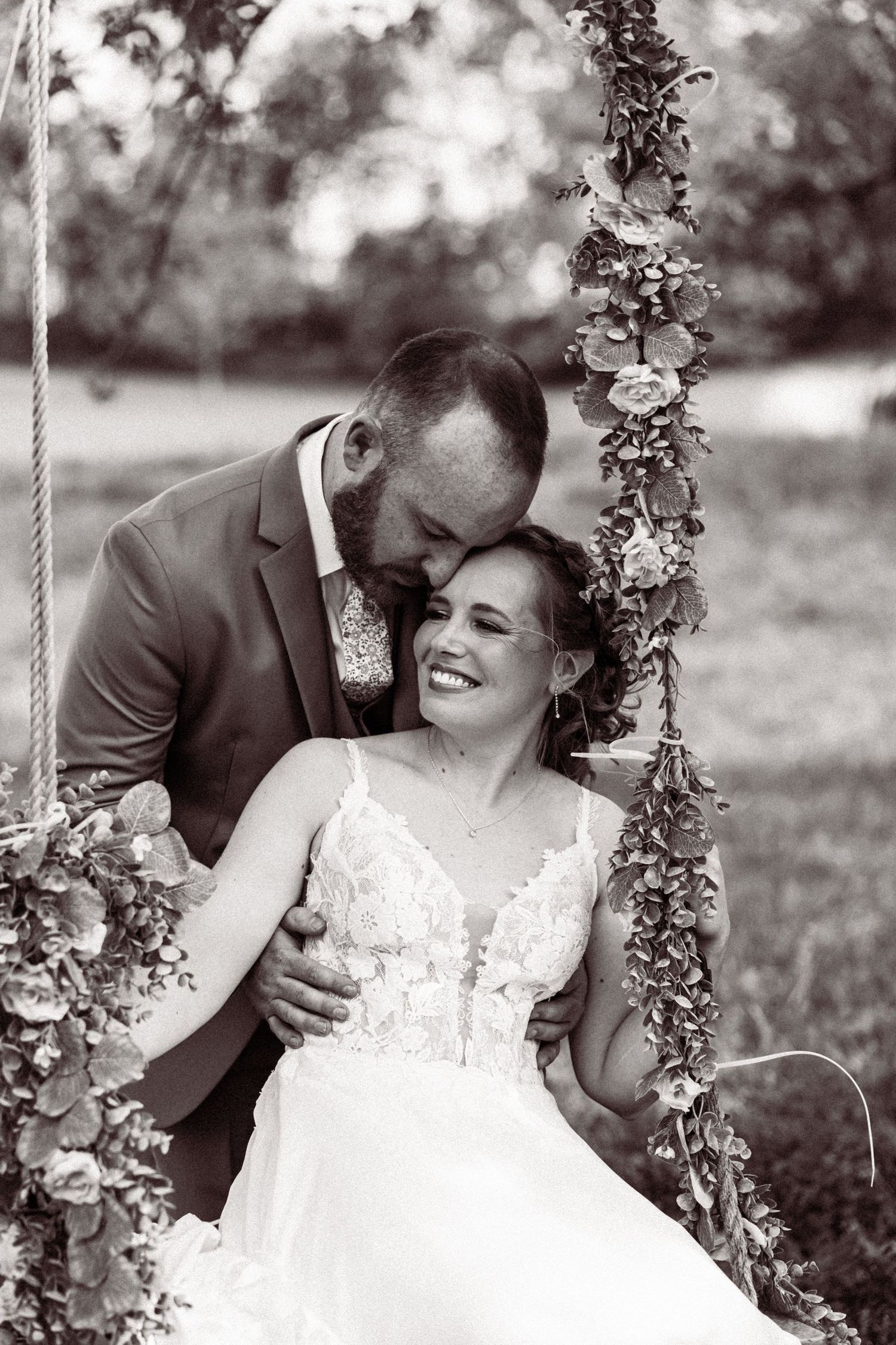 Black and white photo of groom embracing smiling bride on floral-decorated swing outdoors.