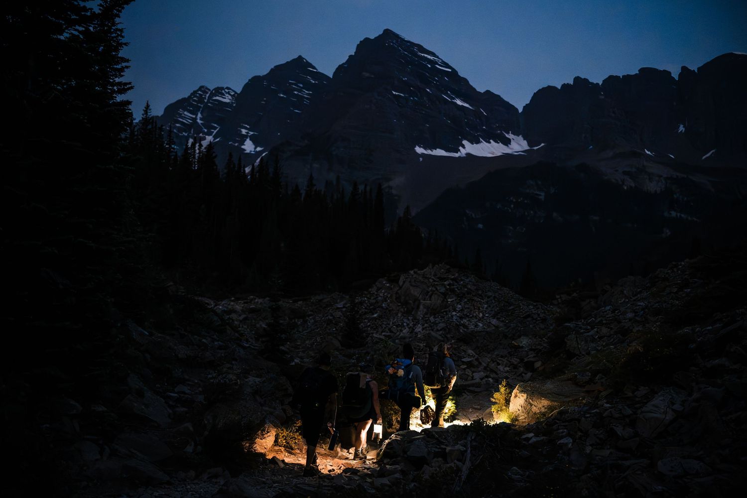 Hikers with glowing lanterns gather at night beneath dramatic snow-capped mountain peaks in a dark wilderness landscape.