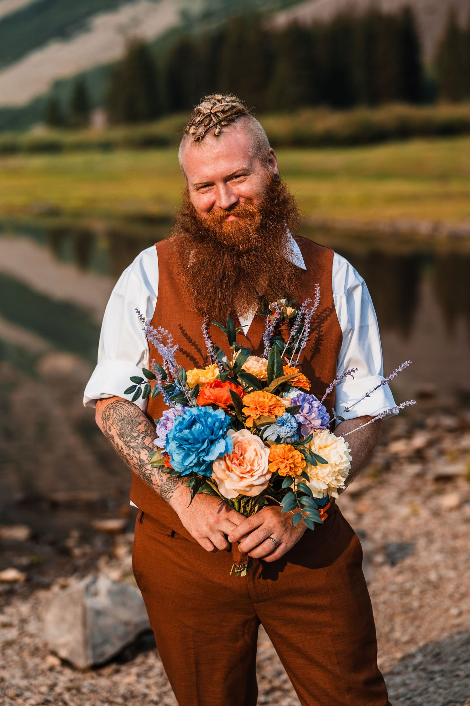 Smiling red-bearded groom in brown vest holds colorful bridal bouquet by a reflective mountain lake.