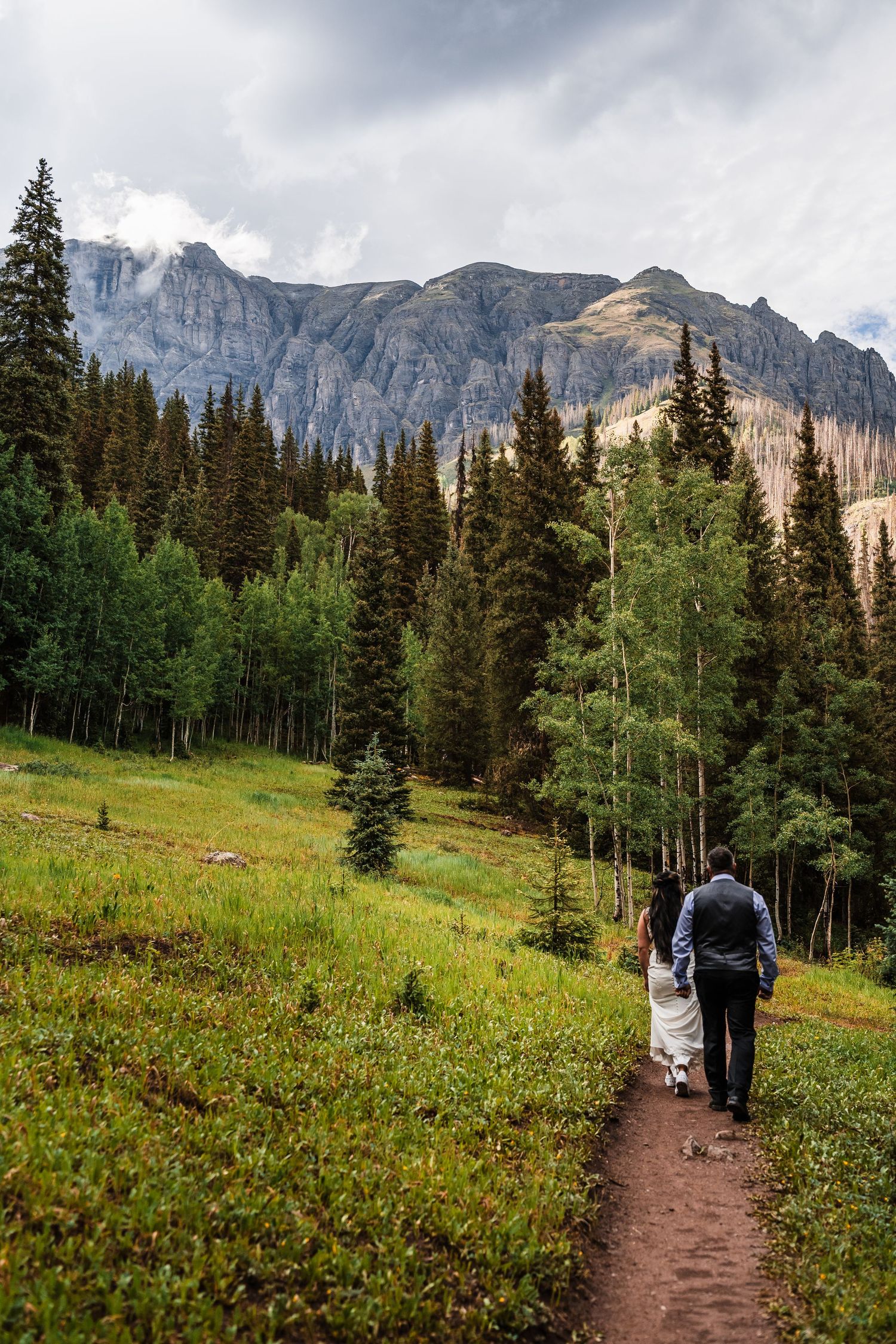 Bride and groom walking hand in hand on a mountain trail with dramatic rocky peaks behind them.