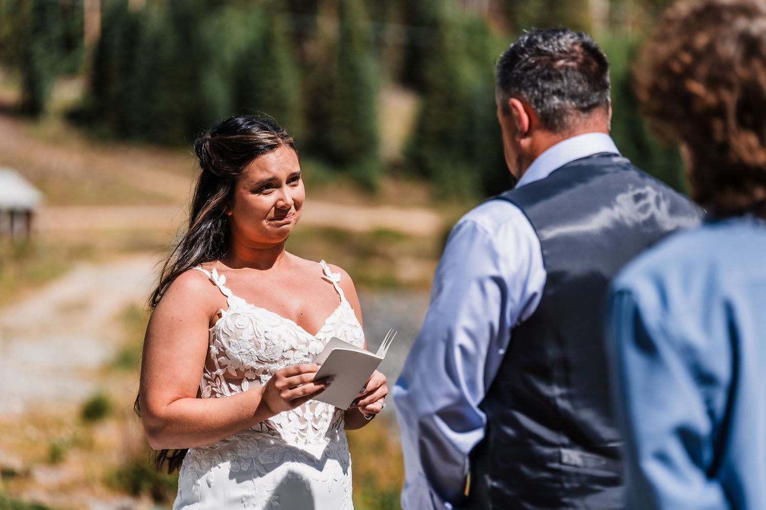 Bride reads wedding vows to groom outdoors in a forest setting, wearing a lace wedding dress.