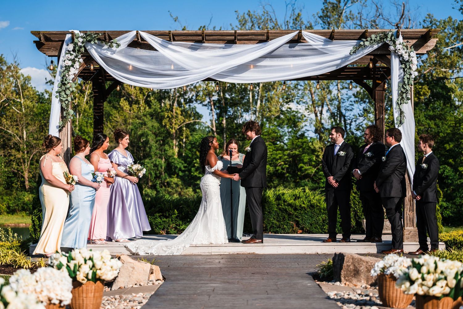 Wedding ceremony under a draped pergola with bridesmaids and groomsmen, surrounded by lush greenery.