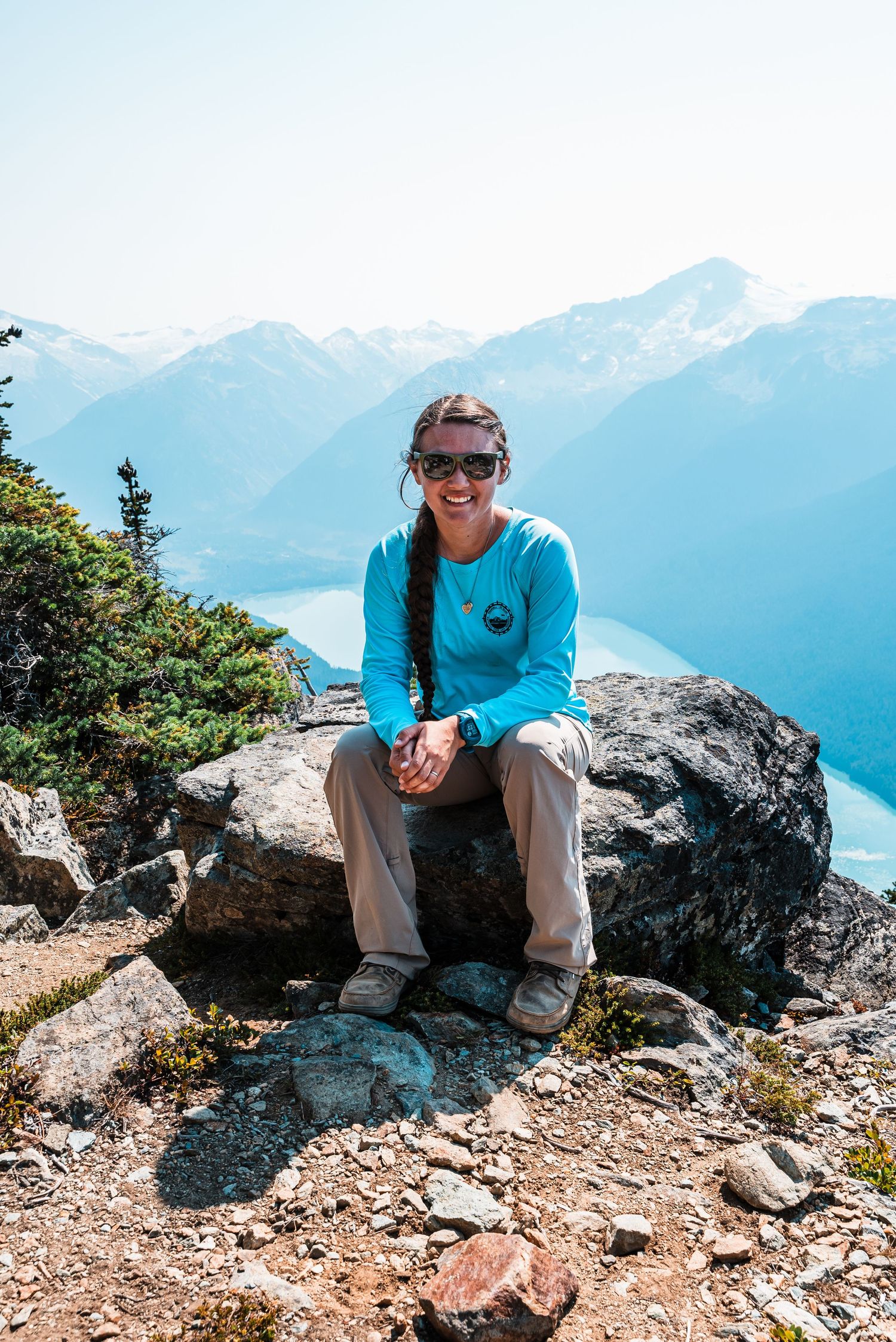 Person in blue shirt sits on rocky mountain overlook with scenic mountain vista.