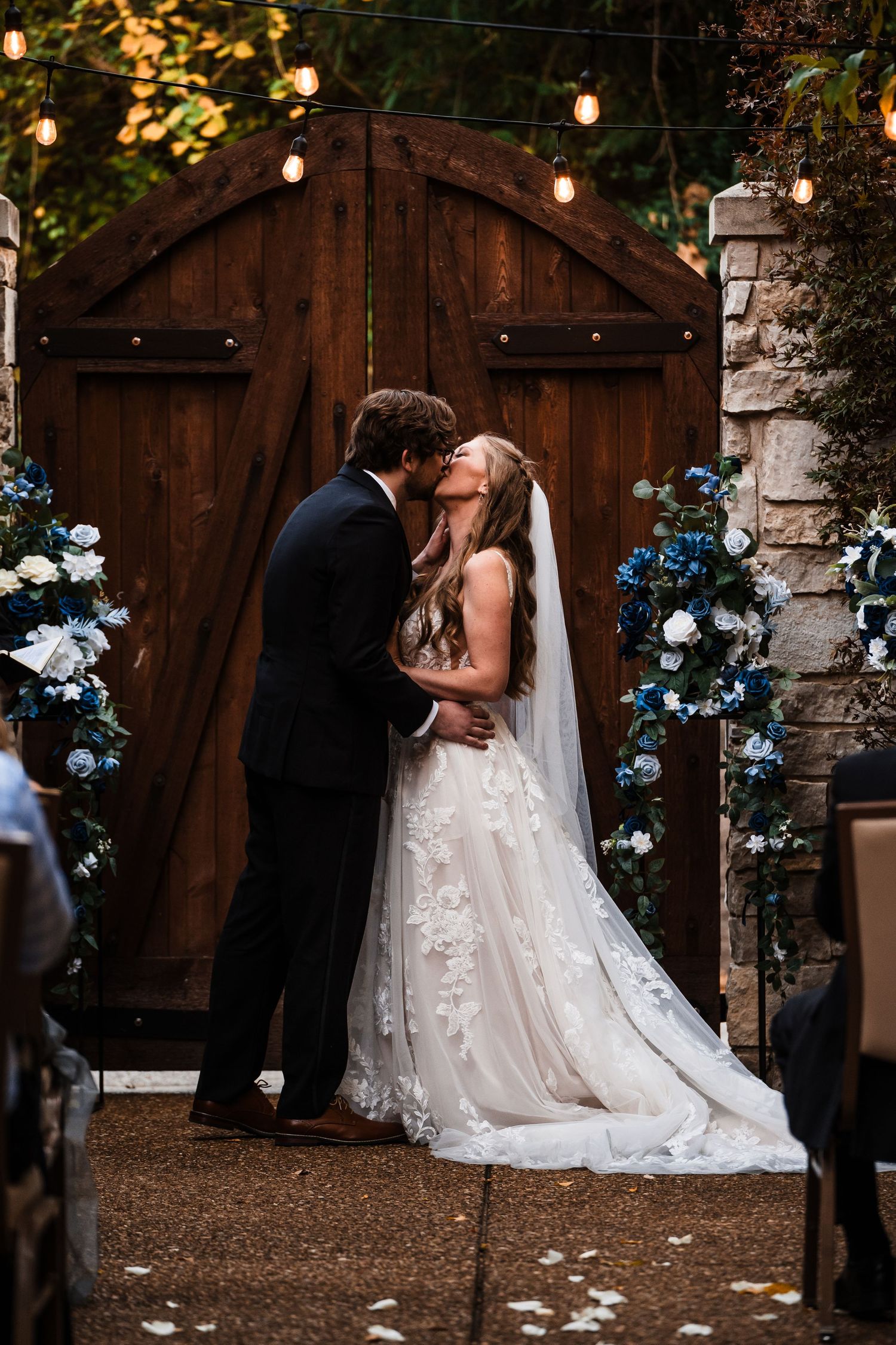 Romantic wedding kiss in front of wooden barn doors adorned with blue flowers and twinkling lights.