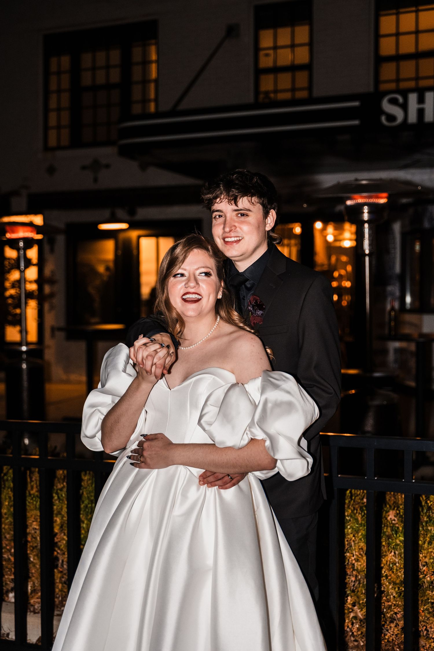 Nighttime portrait of a couple in formal attire outside an illuminated storefront.