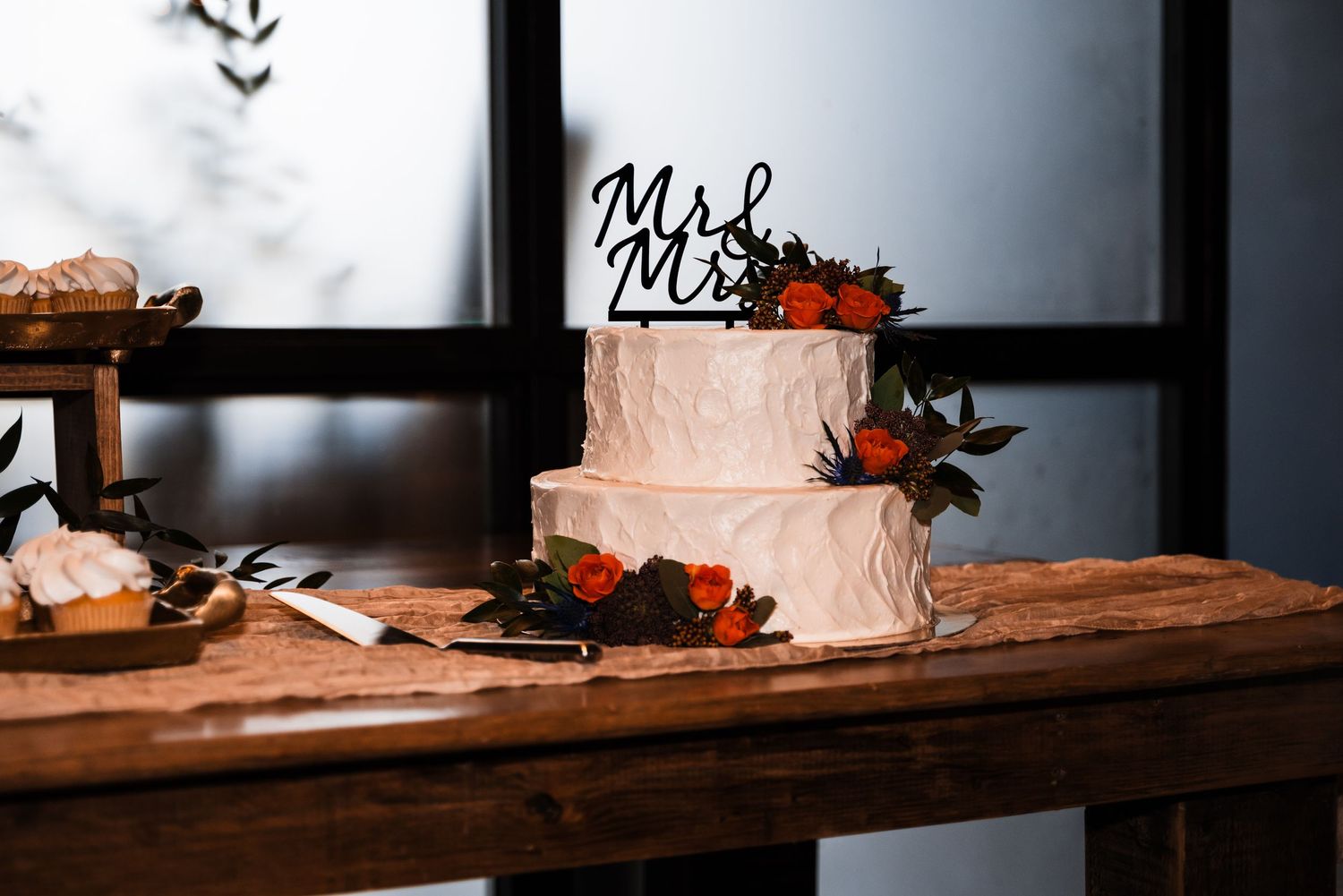 A simple pink wedding cake topped with 'Mr & Mrs' sits on a rustic wooden table.