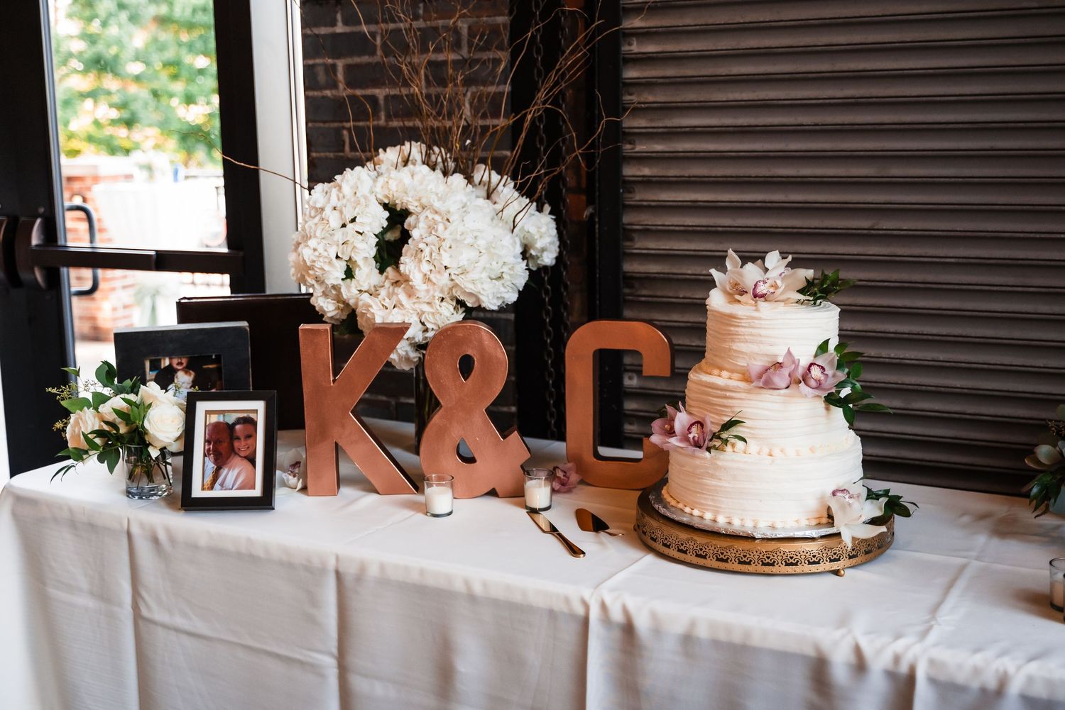 Elegant wedding cake table with a three-tiered white cake, rustic wooden letters K&C, and white floral arrangements.