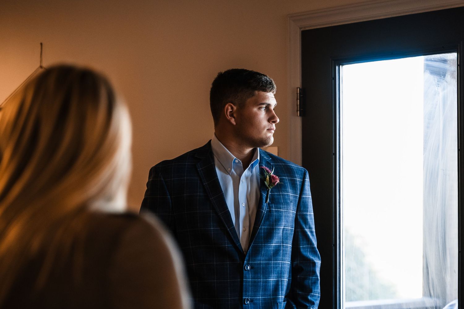 Side profile of someone in a blue checkered suit jacket gazing out a bright window.