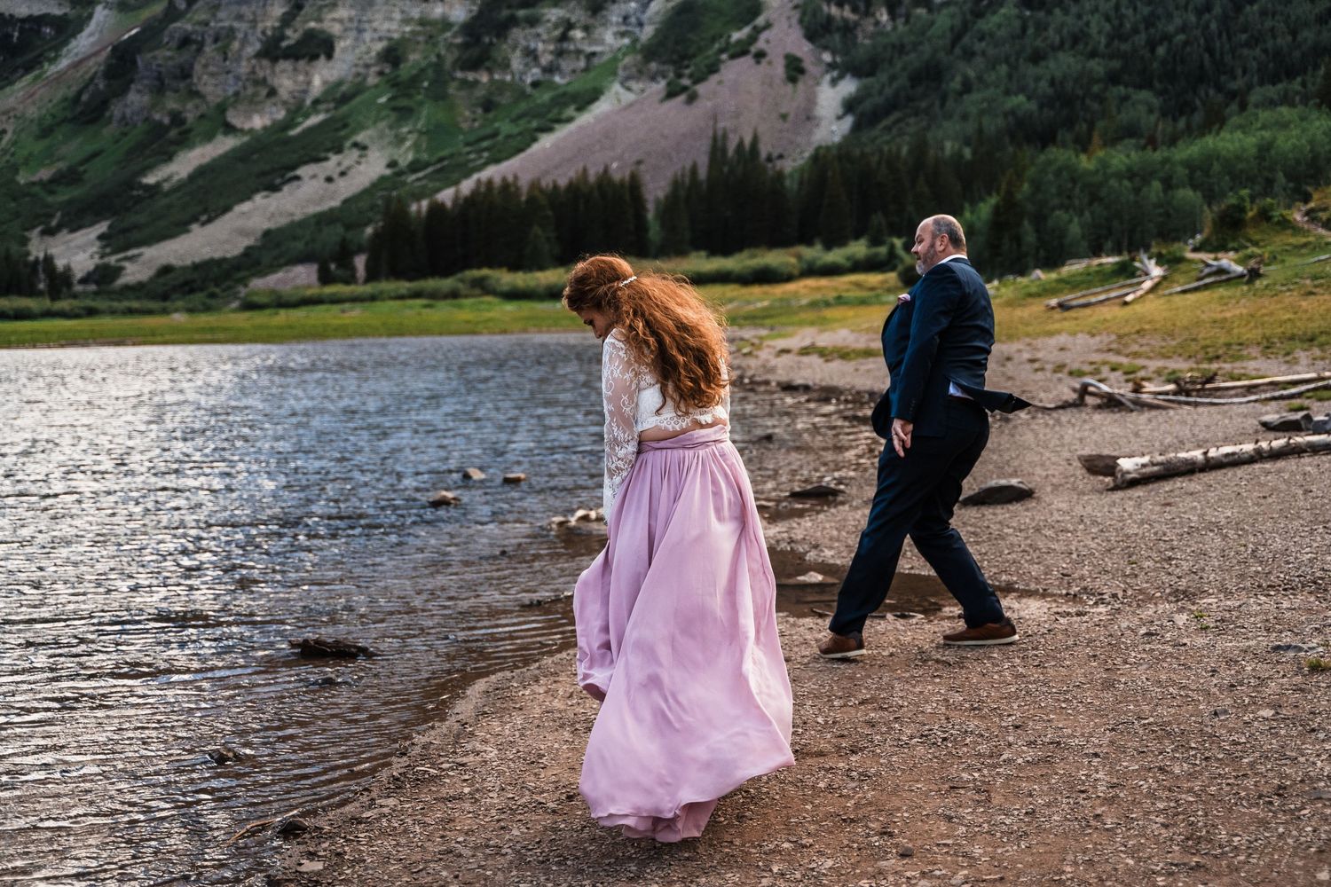 Bride in flowing pink dress walking along scenic mountain shoreline.