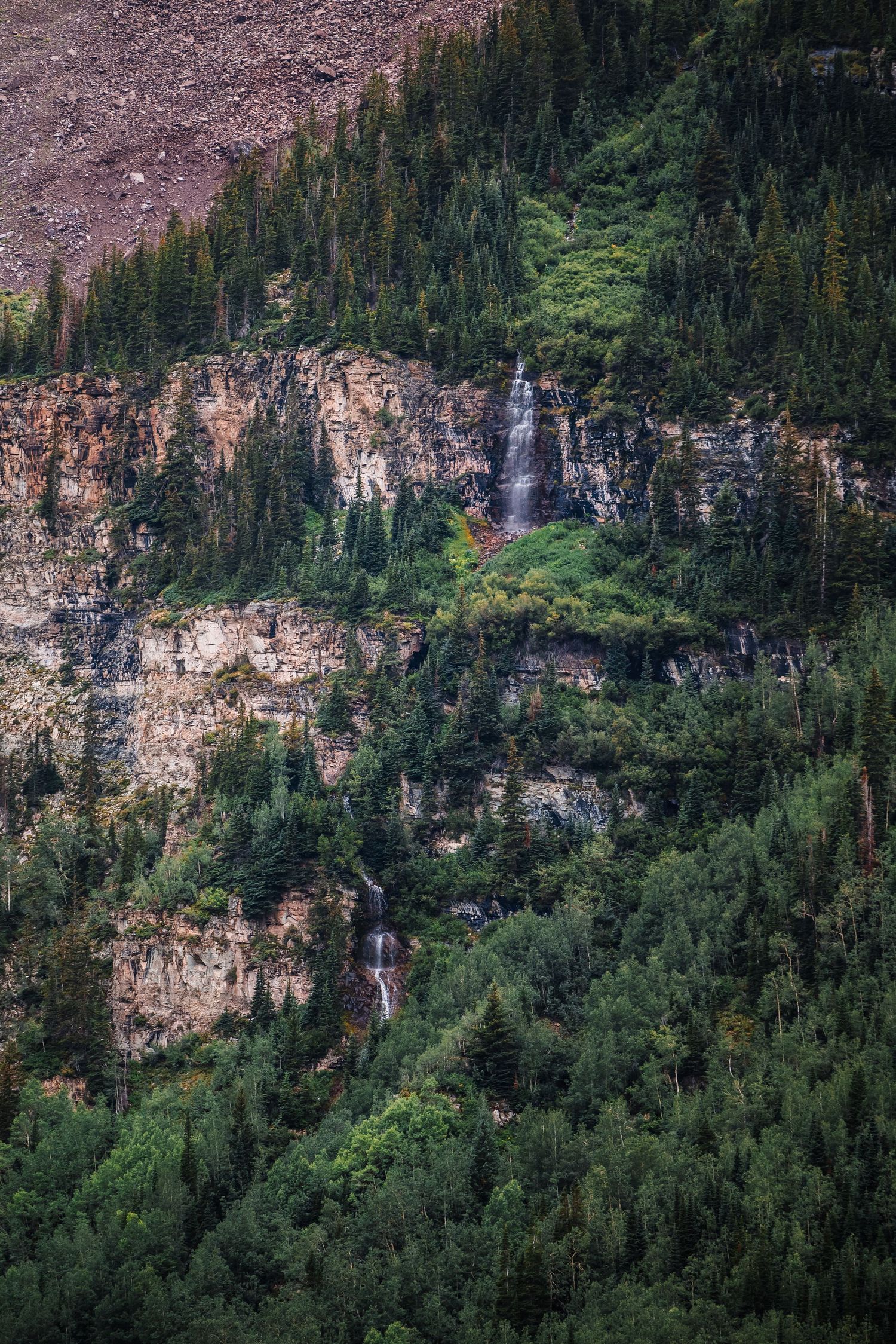 A cascading waterfall flows down rocky cliffs surrounded by lush evergreen forest in the mountains.