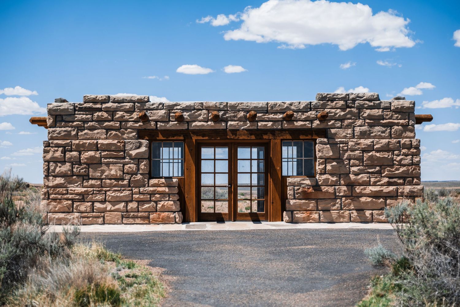 Historic stone cabin with wooden doors and windows stands alone in desert landscape under blue sky with white clouds.
