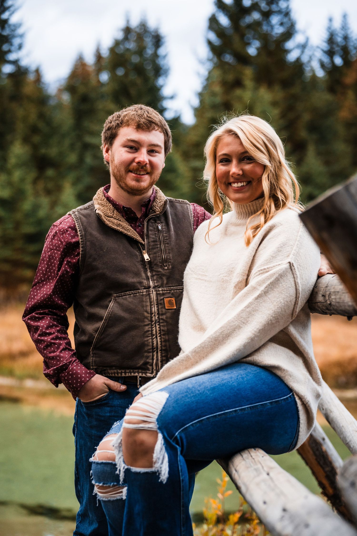 A couple shares a playful moment while sitting on a wooden fence with evergreen trees in the background.