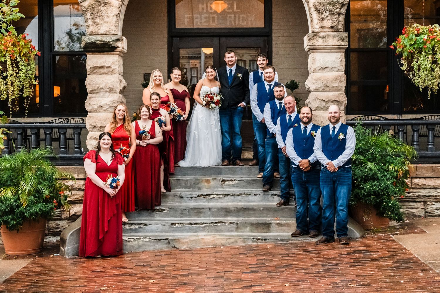 Wedding party in red dresses and blue suits pose on stone steps of historic building entrance.