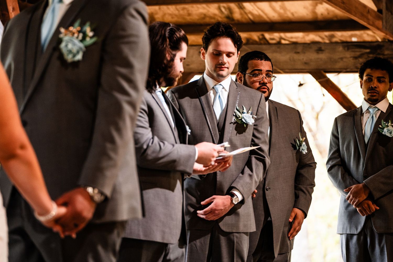 Wedding ceremony takes place under wooden beams with groomsmen in grey suits listening to officiant.