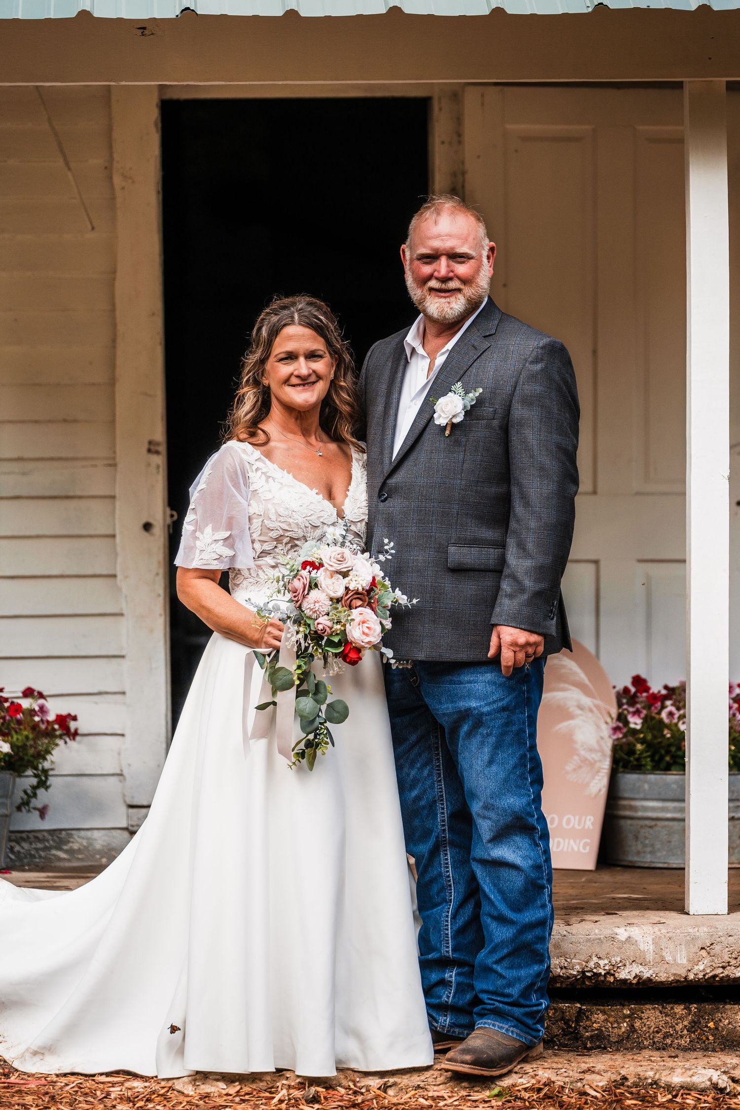 A couple in wedding attire poses together on a rustic white porch during their country-style outdoor wedding celebration.