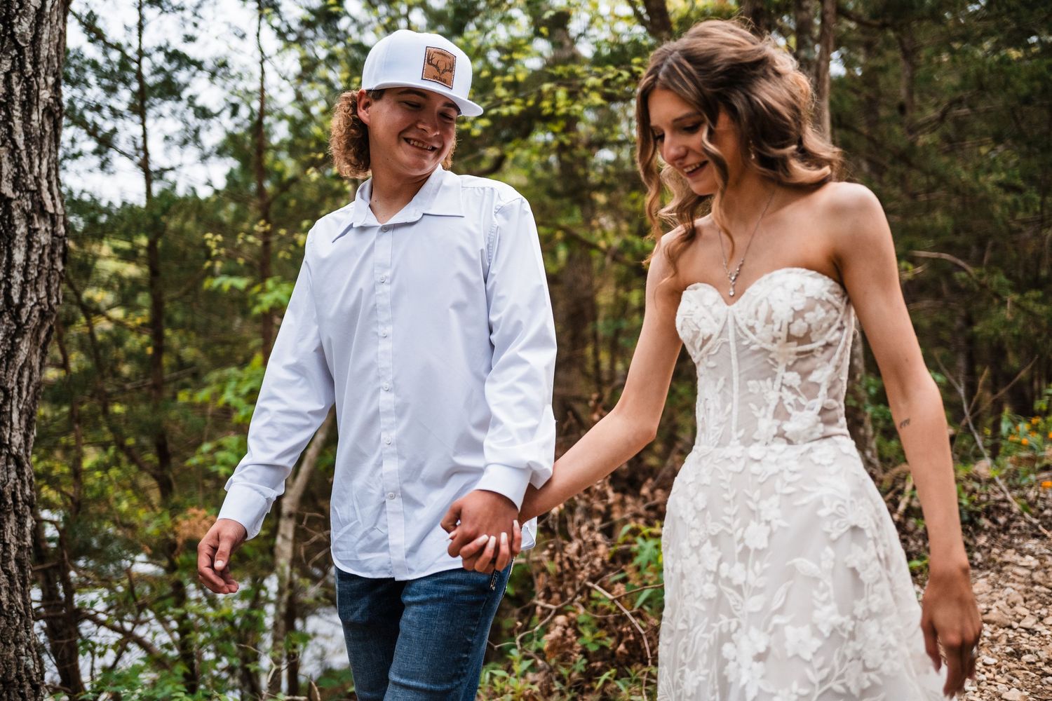 A couple holds hands in nature, with the bride wearing a detailed white lace gown against a forest backdrop.