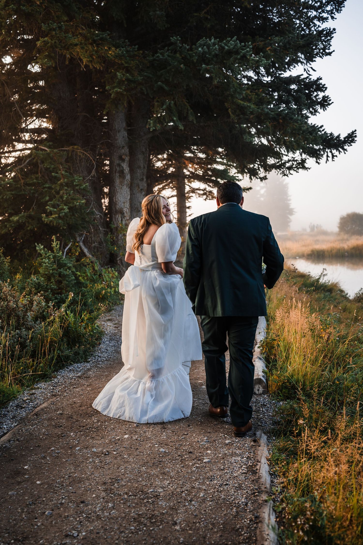A couple in wedding attire walk along a scenic mountain path during golden hour.