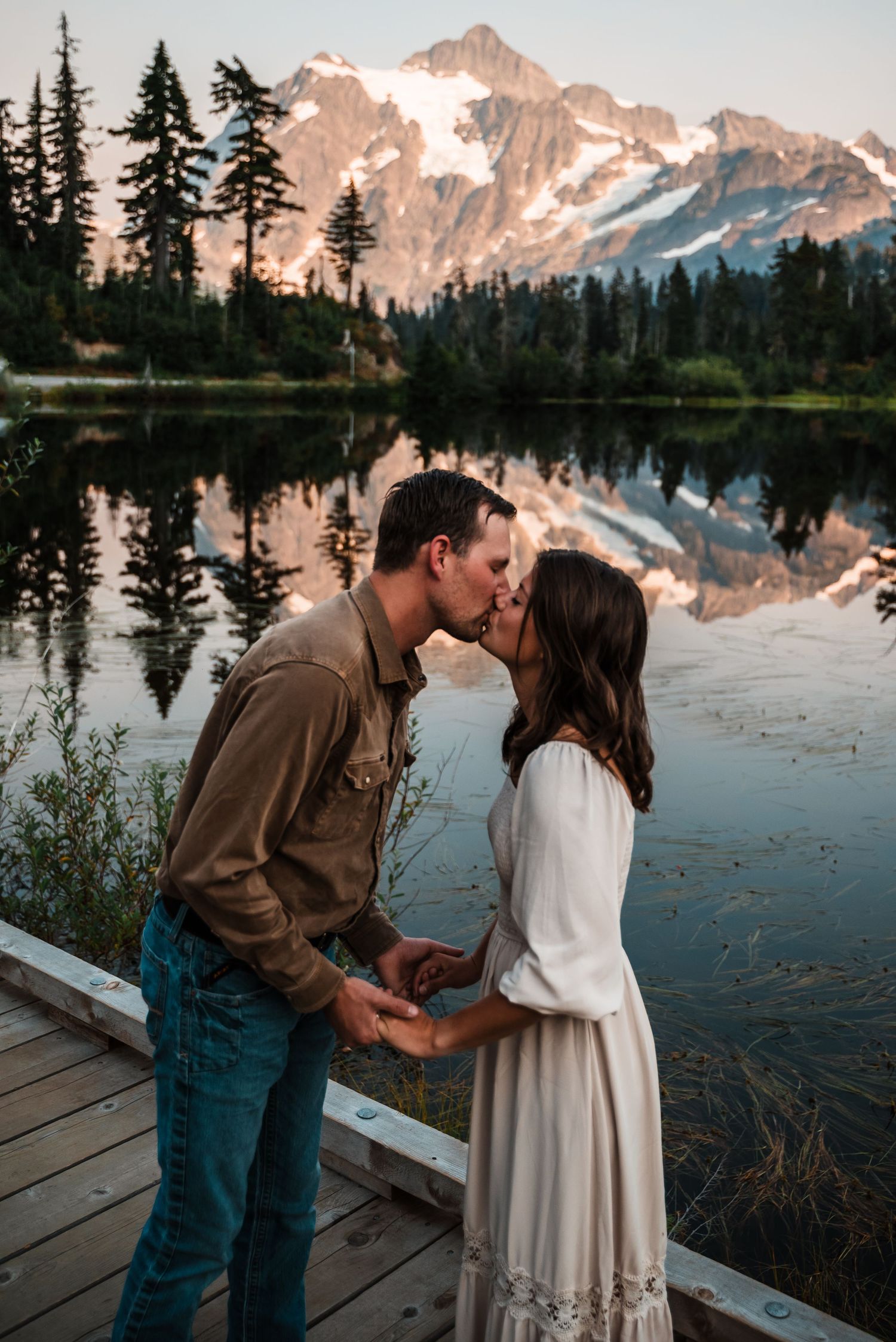 A couple shares a romantic moment on a dock beside a mountain lake with snow-capped peaks reflecting in the calm water.