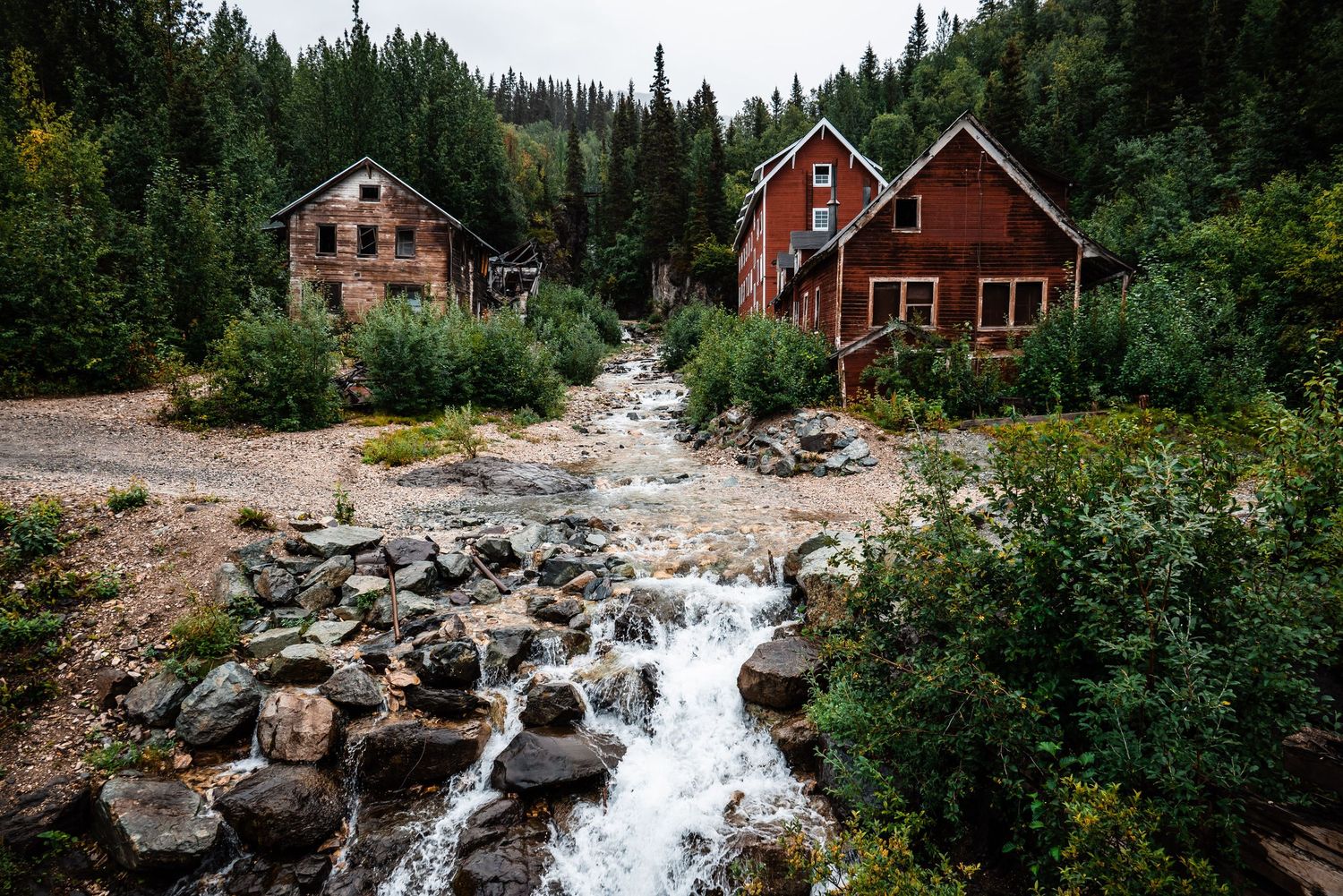 Two red wooden cabins sit alongside a rushing mountain stream surrounded by evergreen forest.