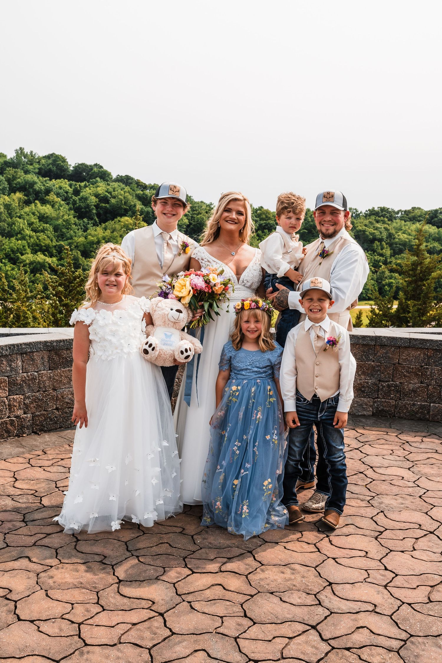 Wedding party of adults and children pose together on stone patio with lush green hills in background.