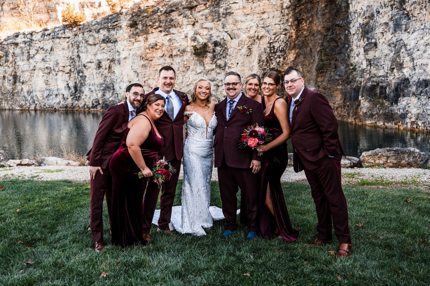 Wedding party poses against a weathered stone wall in burgundy and white attire.