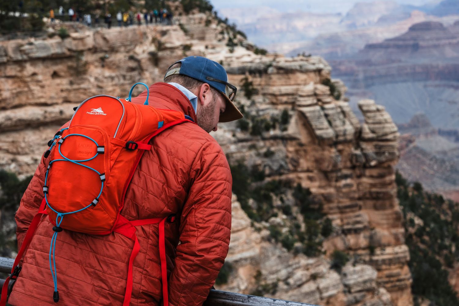 Hiker in orange jacket with backpack overlooking Grand Canyon's majestic rock formations in the distance.