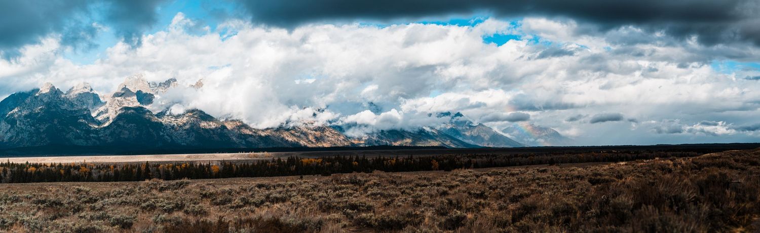 A dramatic panoramic landscape of snow-capped mountains under stormy clouds with dark peaks and rugged terrain.