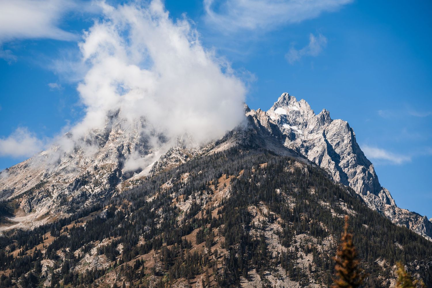 Majestic mountain peak partially shrouded in white clouds against a vibrant blue sky in Grand Teton National Park.