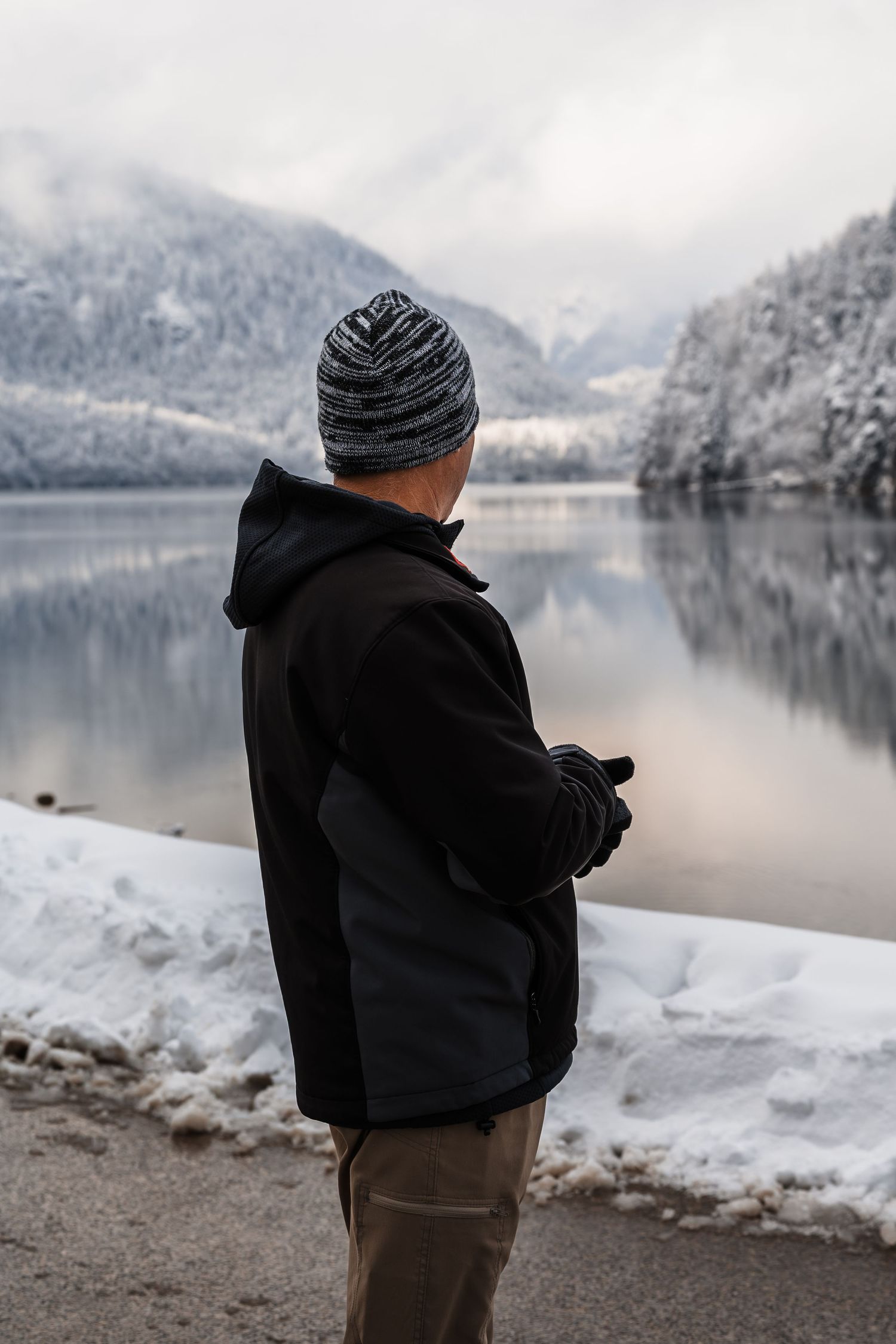 Person in black winter clothing stands at snowy lake shore with mountains and reflective water in winter landscape.