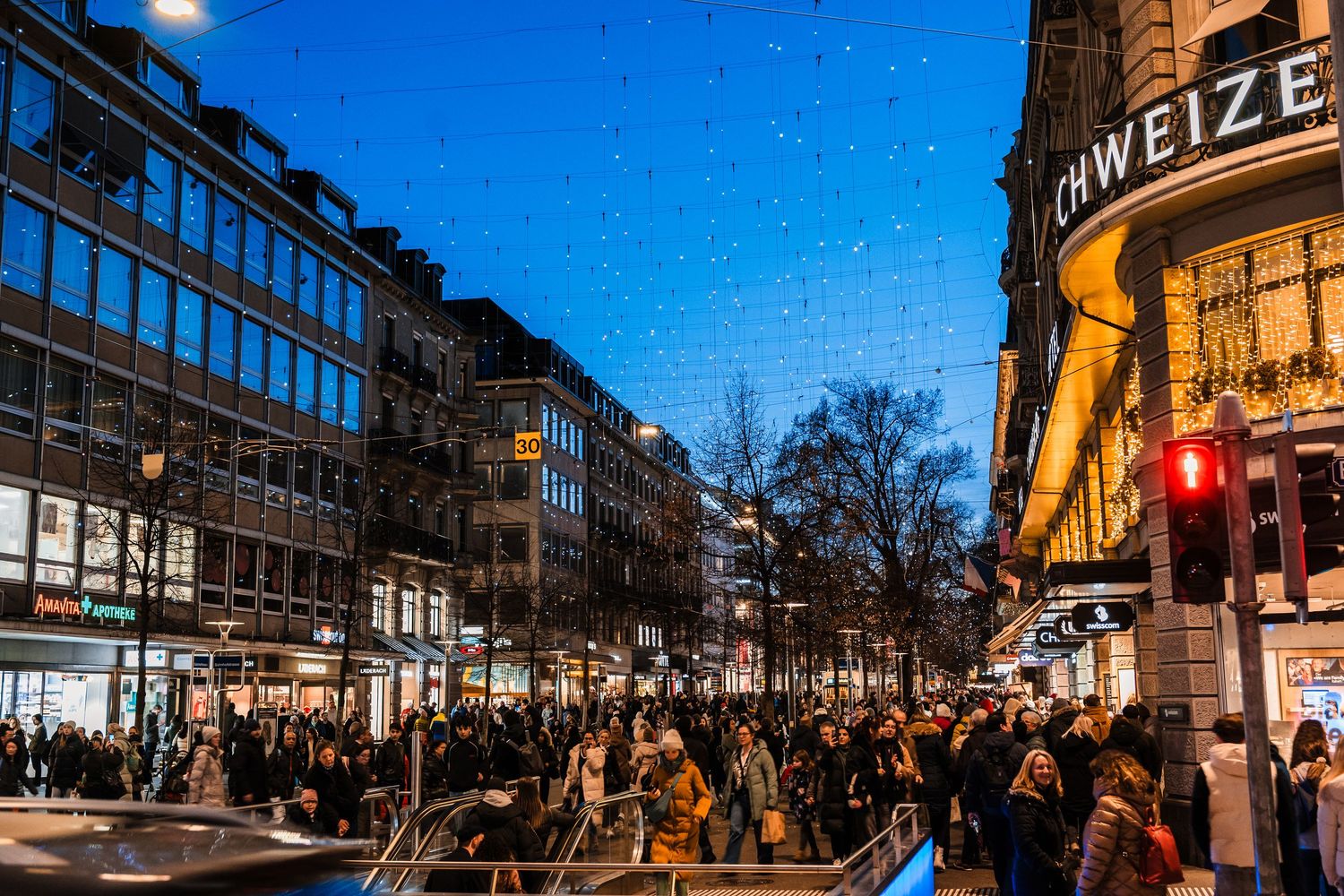 Panoramic view of a bustling city street at dusk with illuminated storefronts and pedestrians.