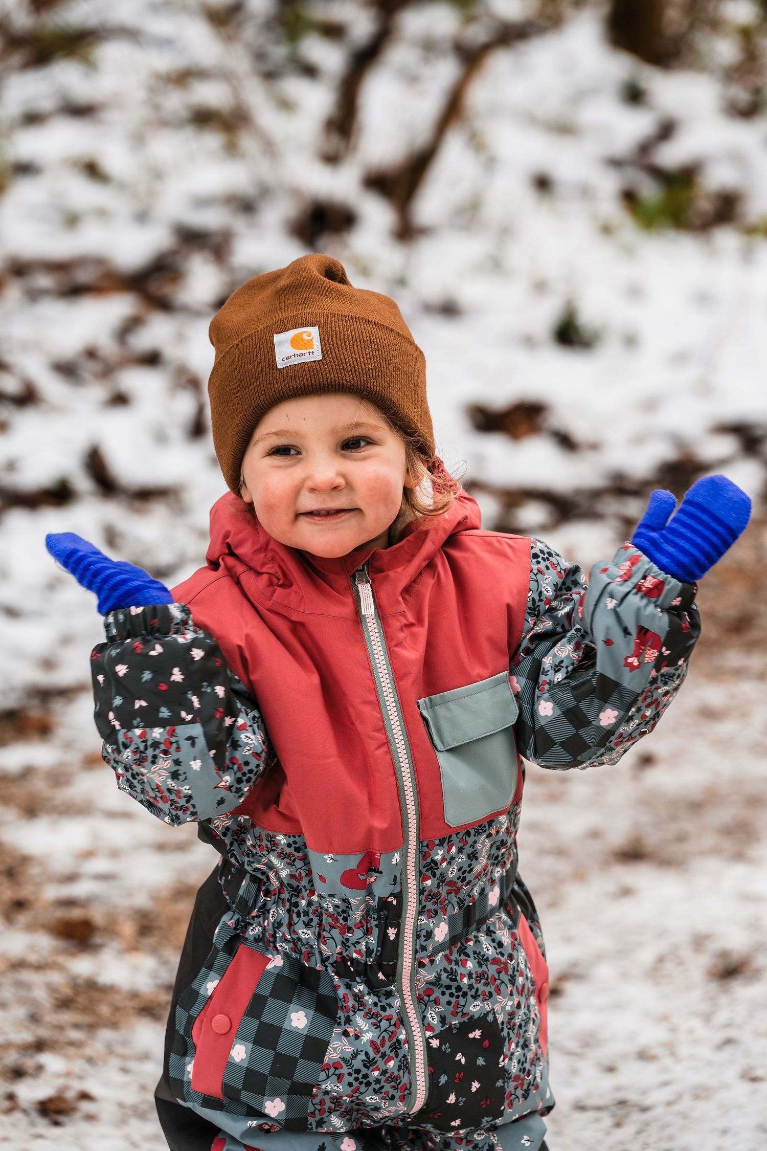 Small child in winter clothing smiles and raises arms while playing in snowy outdoor setting.